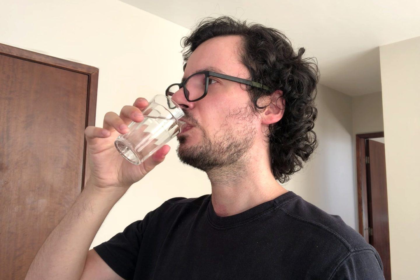 Homem de cabelo ondulado, barba, óculos e camiseta preta, bebendo água em um copo americano.