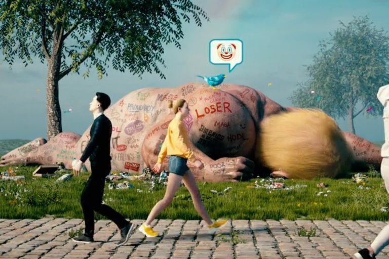 Quadro do vídeo CROSSROADS, de Beeple. Ao fundo, uma pessoa enorme que lembra Donald Trump está deitada, com um pássaro azul do Twitter sobre ela e um balão de conversa com o emoji de um palhaço. Na calçada, pessoas caminham, ignorando o enorme Trump deitado no gramado atrás.