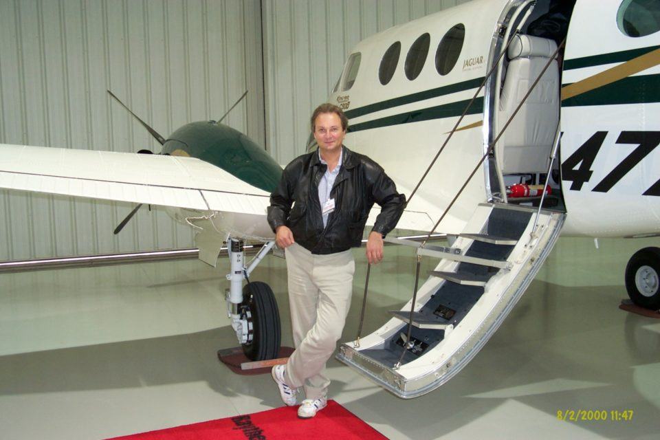 Homem branco, de calça sarja e jaqueta marrom, com o braço apoiado no corrimão das escadas de um avião de pequeno porte, dentro de um galpão.