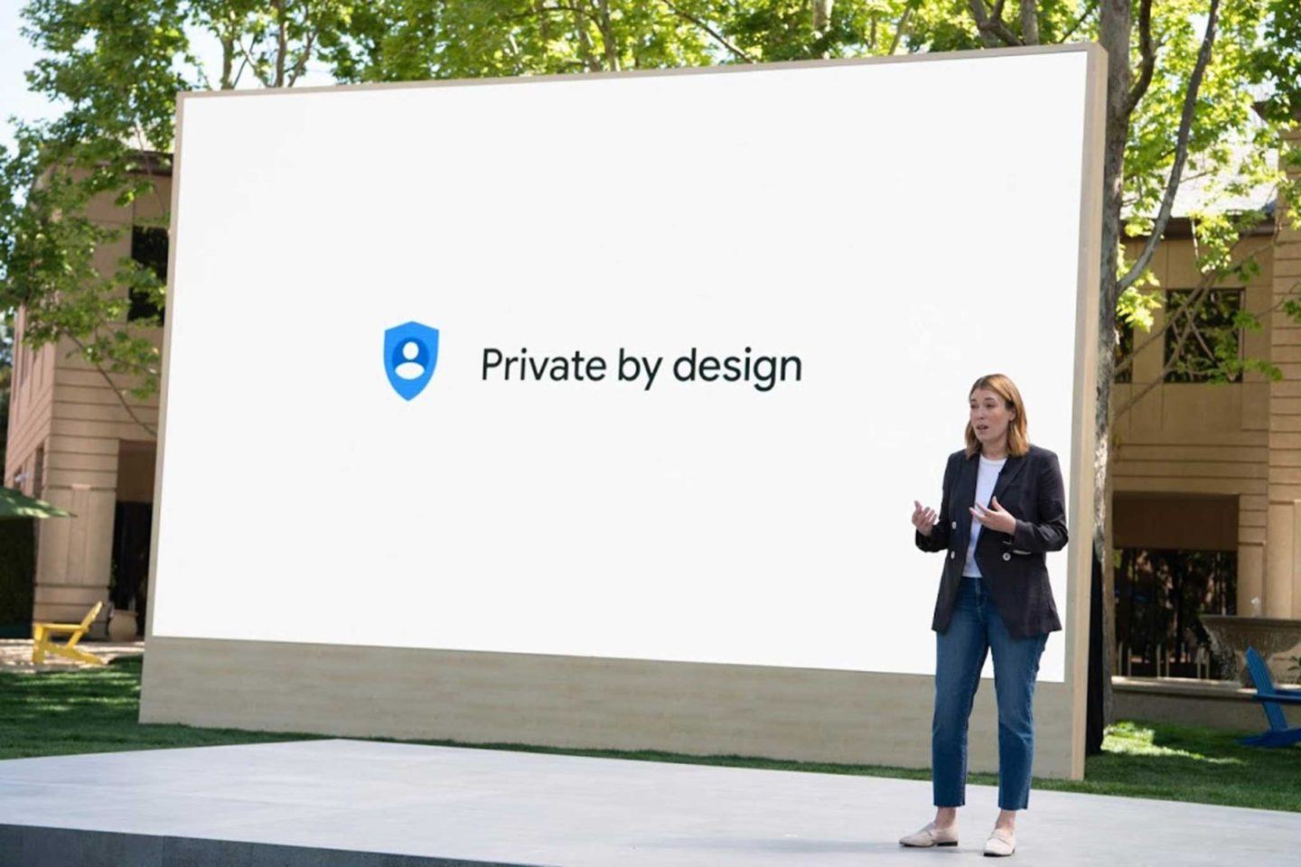 Mulher de calça jeans e casaquinho, em um ambiente ao ar livre com árvores, e ao fundo uma enorme tela com os dizeres, em inglês, "Priority by design".