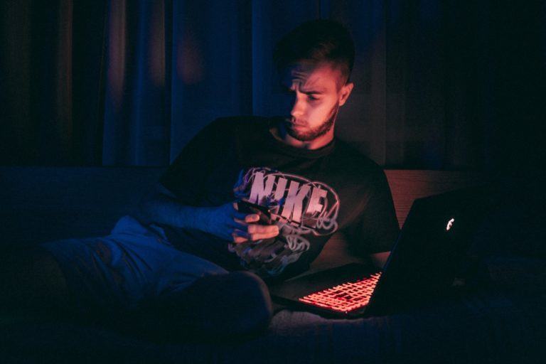 Homem branco de camiseta preta deitado em um sofá, olhando para o celular, iluminado pelas telas do celular e notebook.