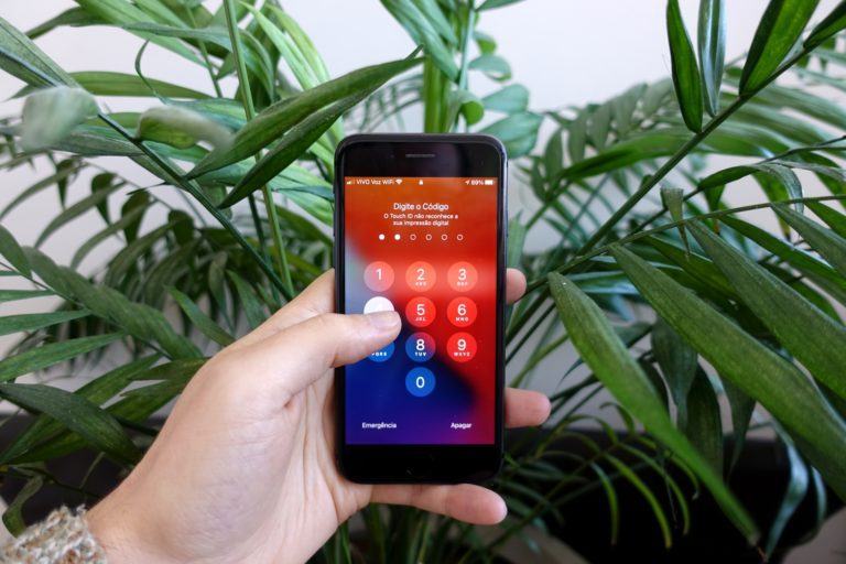 Mão segurando um iPhone com a tela pedindo o código de desbloqueio. Ao fundo, folhas de uma palmeira.