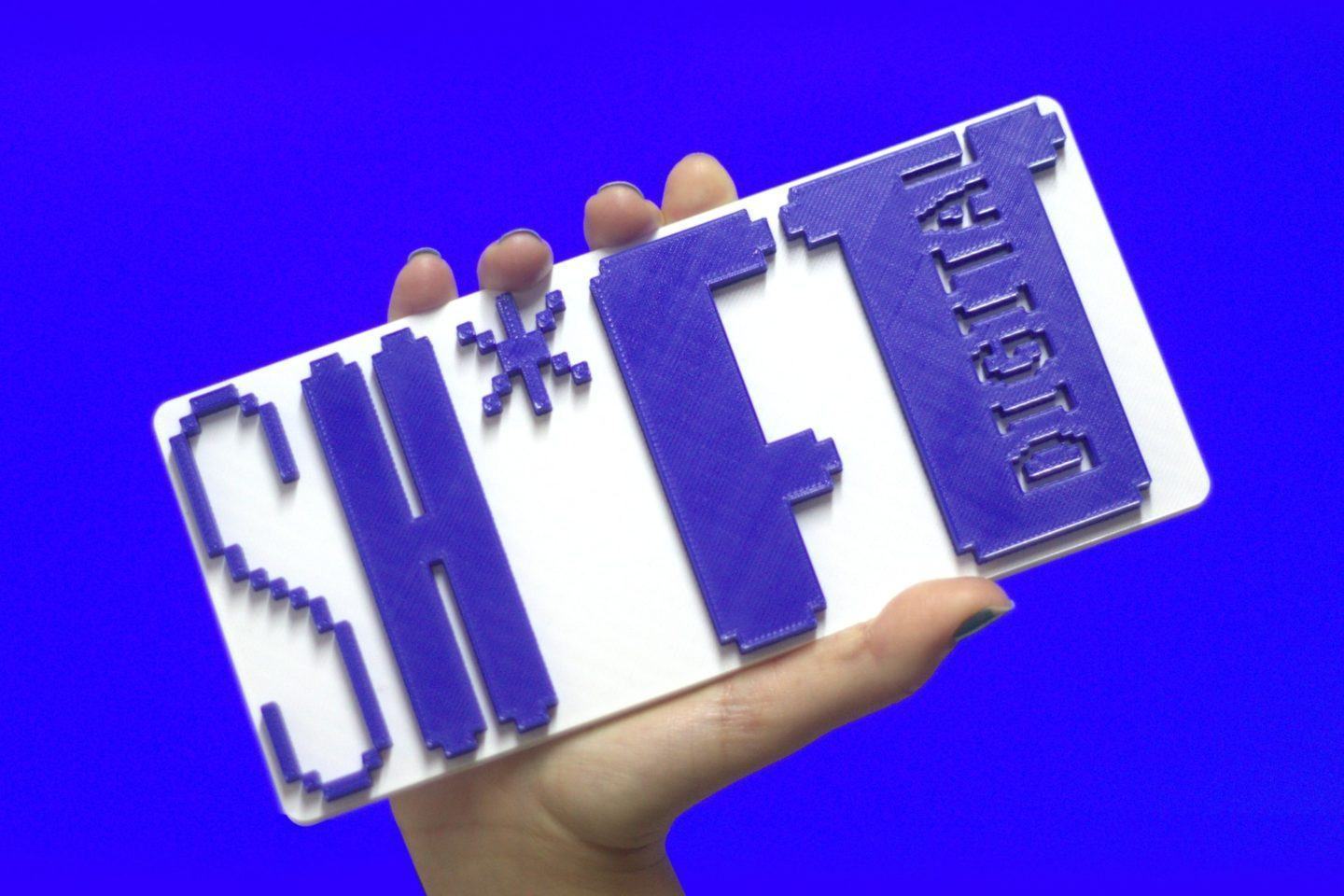 Mão segurando um letreiro escrito “SH*FT Digital” contra um fundo azul.