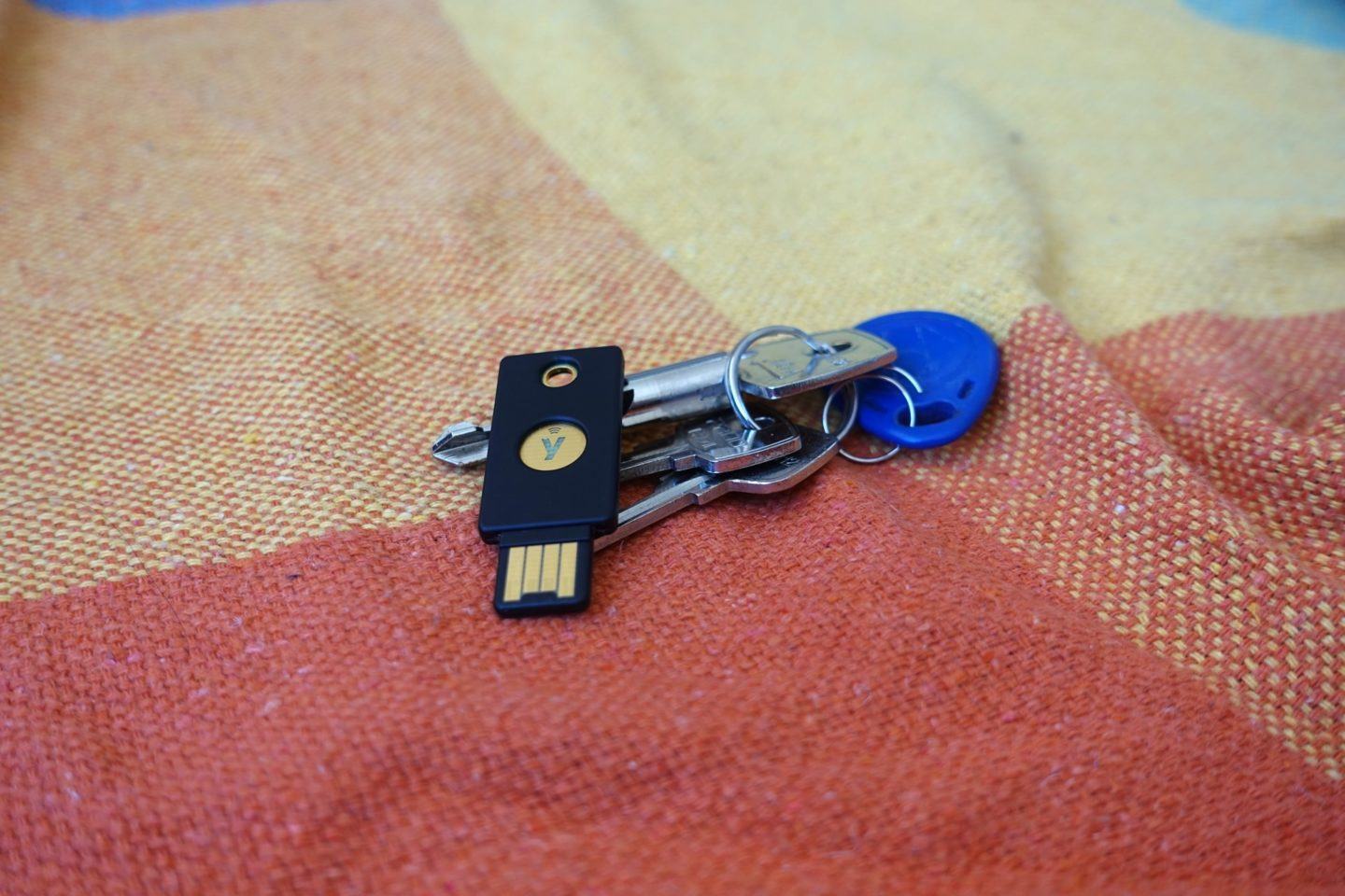 Uma YubiKey 5 NFC junto a um molho de chaves comuns, em cima de um tecido grosso e colorido (amarelo, vermelho e laranja).