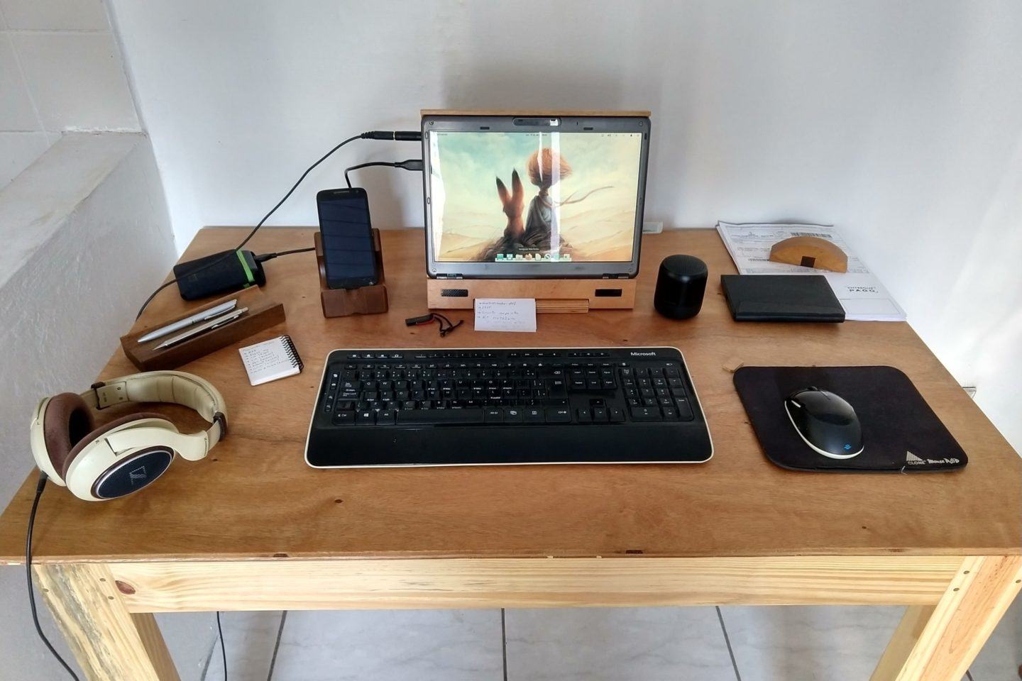 Vista frontal da mesa de trabalho em casa do Diego, com computador e acessórios espalhados.