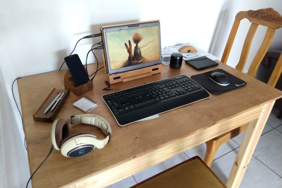 Mesa de madeira, com teclado, mouse e um monitor/notebook desmontado e remontado em um suporte de madeira. Ao redor, acessórios como fones de ouvido e um e-reader.