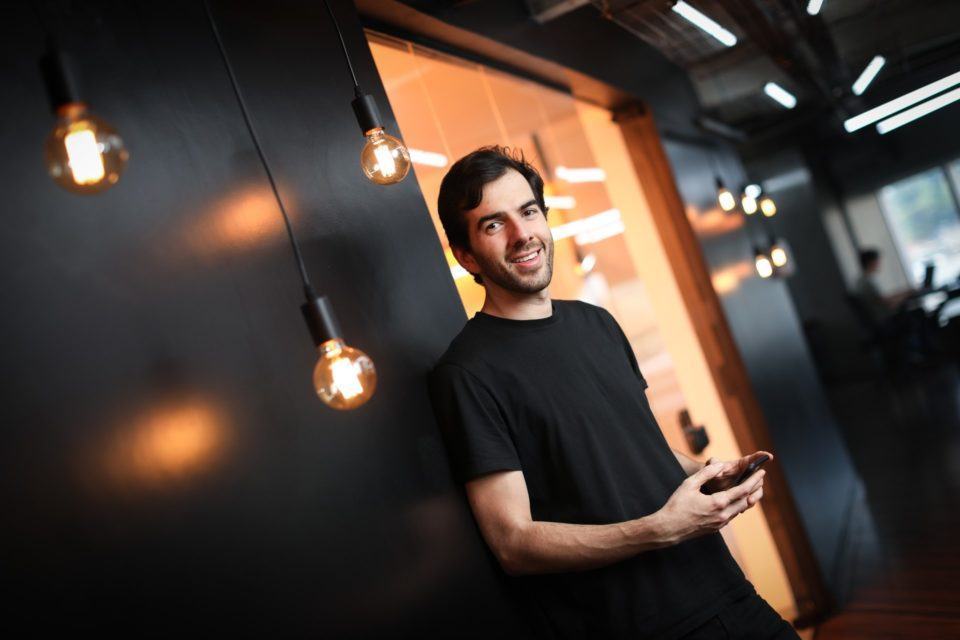 Homem branco segurando um celular, encostado em uma parede preta com lâmpadas de cores quentes penduradas ao redor.