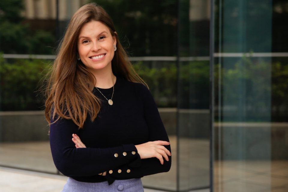 Mulher branca, de cabelos compridos e castanhos, com os braços cruzados, sorrindo, ao lado de uma estrutura de vidro.