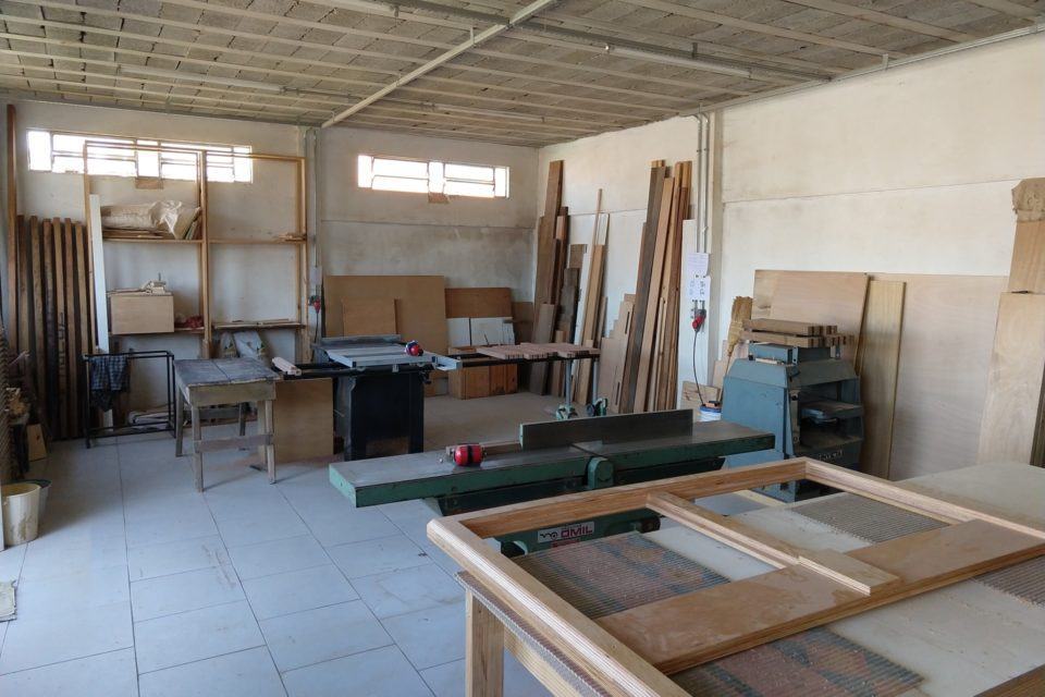 Foto da marcenaria do ângulo contrário, com mais mesas e máquinas e janelas estreitas próximas ao teto.