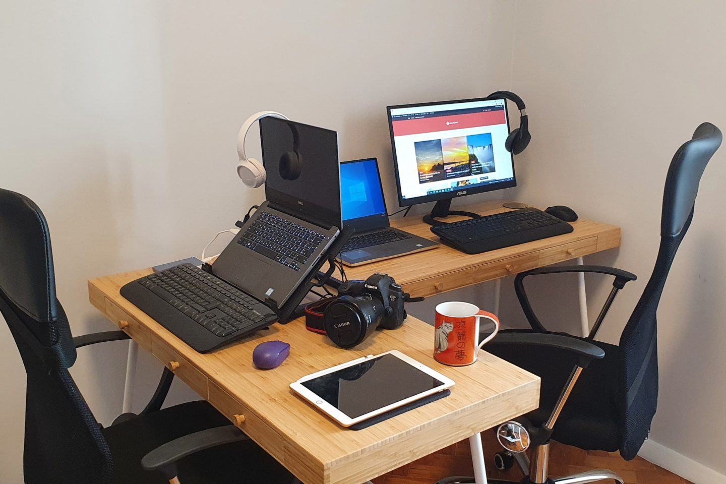 Duas mesas em “L”, com notebooks, teclados, mouses, uma câmera, um tablet e uma caneca sobre elas.