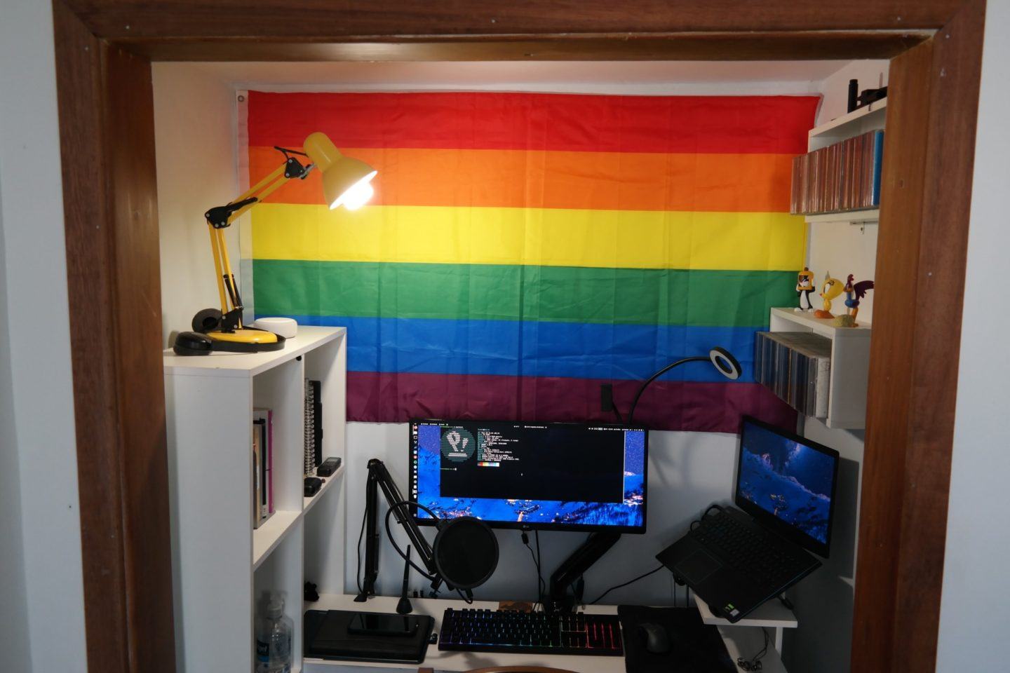 Mesa de trabalho com uma tela e um computador, nichos nas laterais e, ao fundo, uma bandeira LGBT.