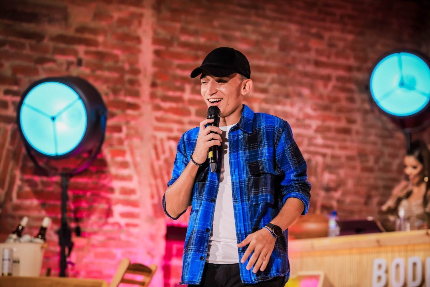 Homem branco, de boné preto e camisa xadrez azul, segura um microfone enquanto canta em um ambiente com iluminação colorida (azul e vermelha) e paredes de tijolinhos atrás.