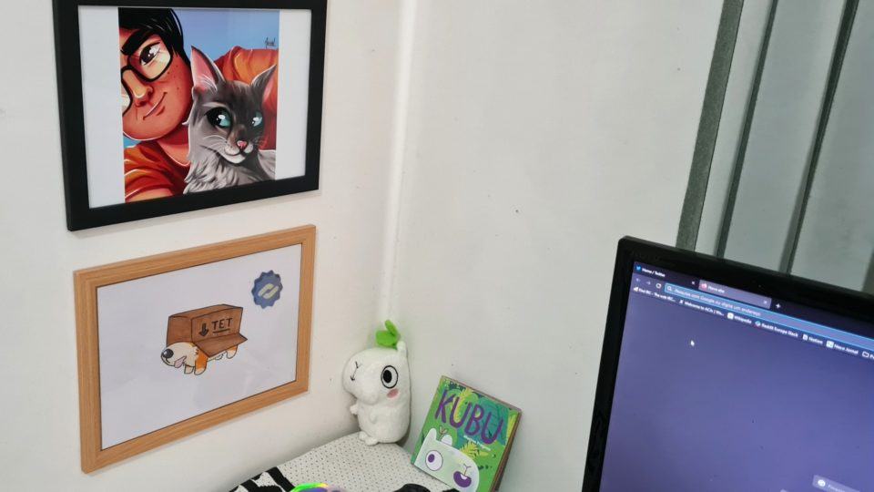 Detalhe do canto do escritório do Tet, mostrando dois quadros de desenhos/avatares dele, uma pelúcia e um livro da pelúcia. No lado direito, parte de um monitor.