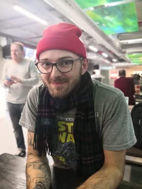 Homem branco, de barba, óculos de grau, toca rosa, camiseta cinza de Star Wars e cachecol preto, olhando para a câmera em uma espécie de aeroporto ou estação de metrô (fundo desfocado).