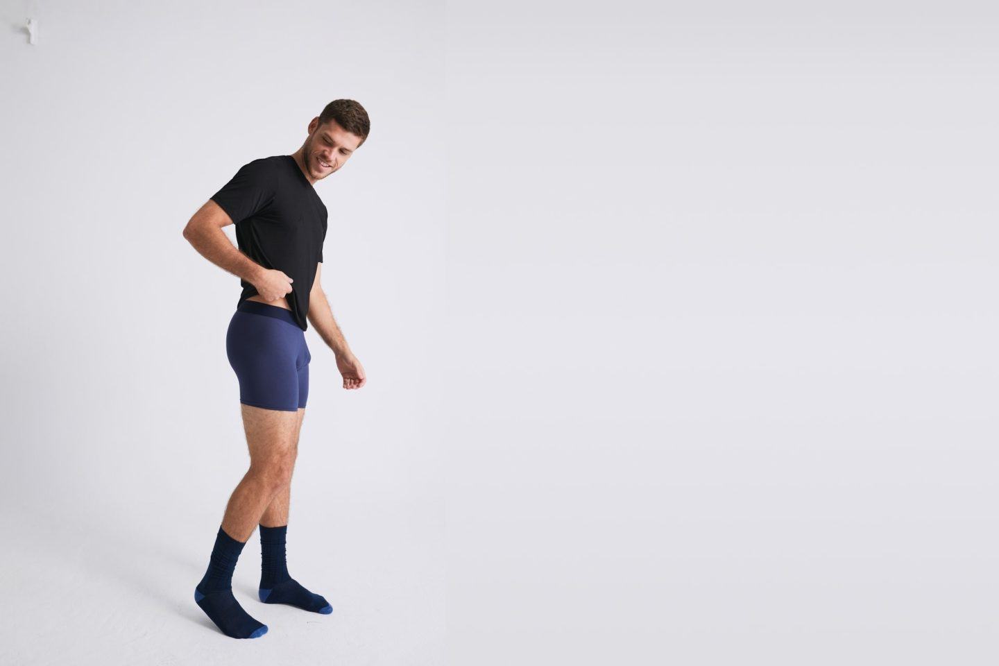 Homem branco, de meias e camiseta preta e cueca azul, em um ambiente amplo e todo branco.