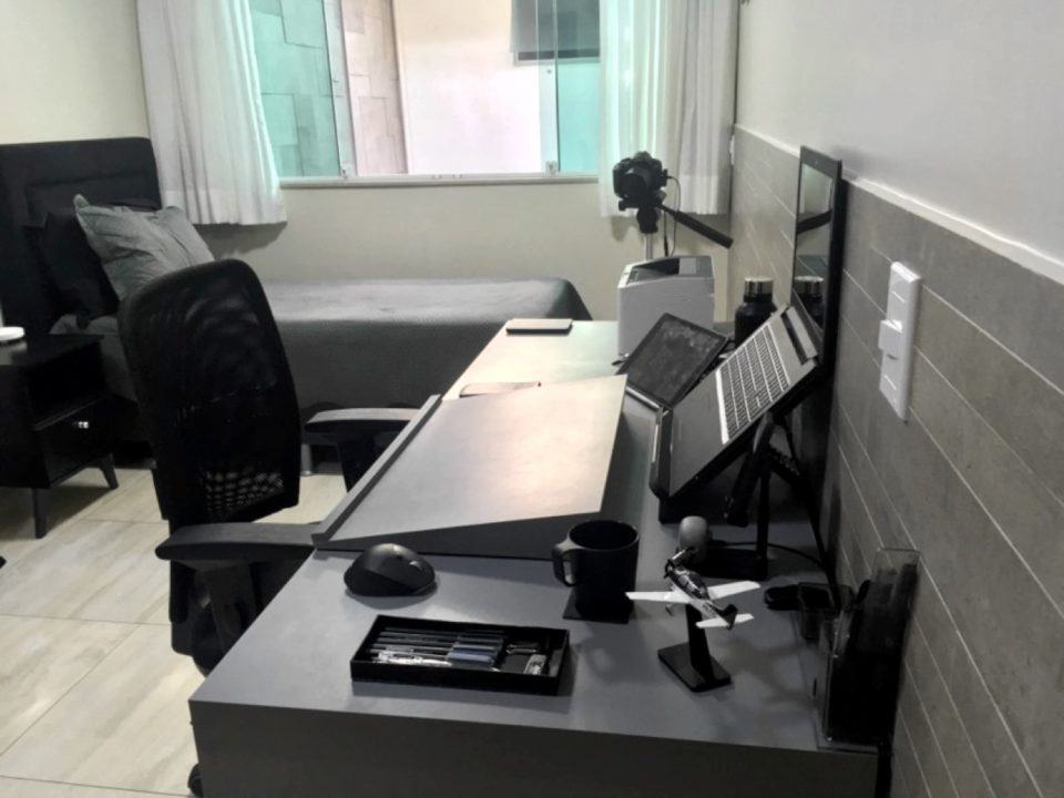 Mesa de trabalho do Pedro Henrique vista por outro ângulo, com a cama e uma janela ao fundo.