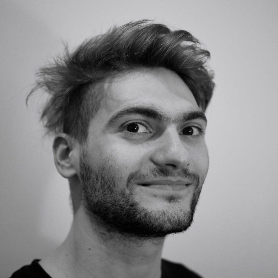 Foto em preto e branco, de homem branco, com barba curta e cabelos espetados, olhando para a câmera.