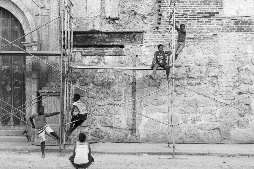 Foto em preto e branco. Cinco crianças em um andaime simples, contra uma parede de pedra e uma porta à esquerda.