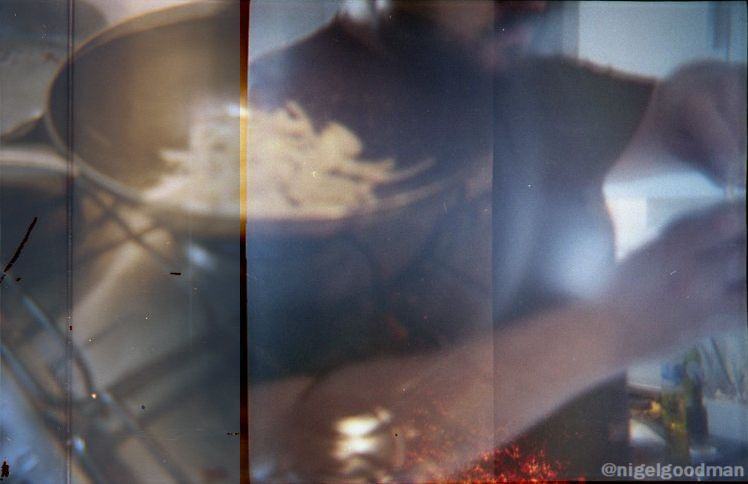 Foto com dupla exposição, mostrando uma panela em um fogão e um homem com barba e camiseta escura.