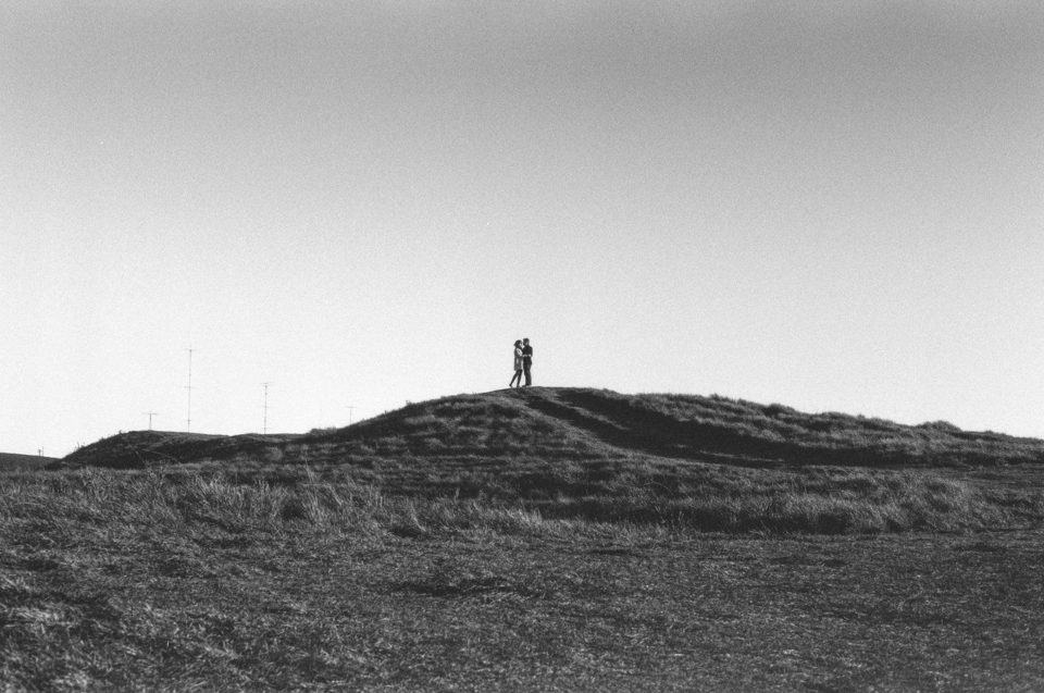 Foto em preto e branco de um casal, à distância, no topo de um rochedo.