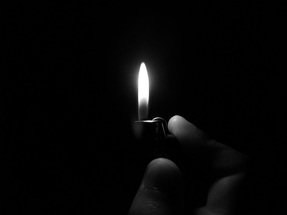 Isqueiro, iluminando a mão que o segura, em um ambiente totalmente escuro.