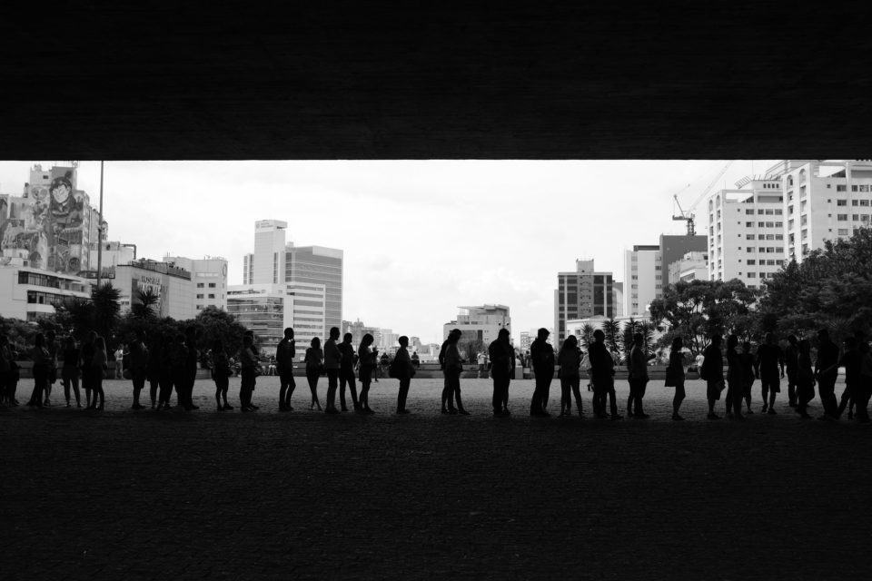 Silhuetas de dezenas de pessoas em uma fila, a média distância, no vão do MASP. Foto em preto e branco.