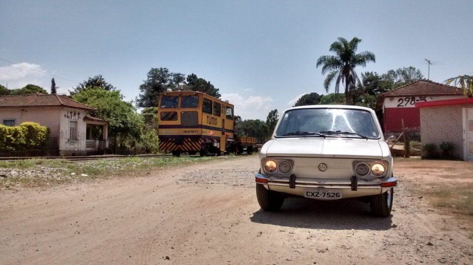 Foto de uma estrada de terra batida, com uma Brasília branca em primeiro plano e algumas casinhas ao fundo.