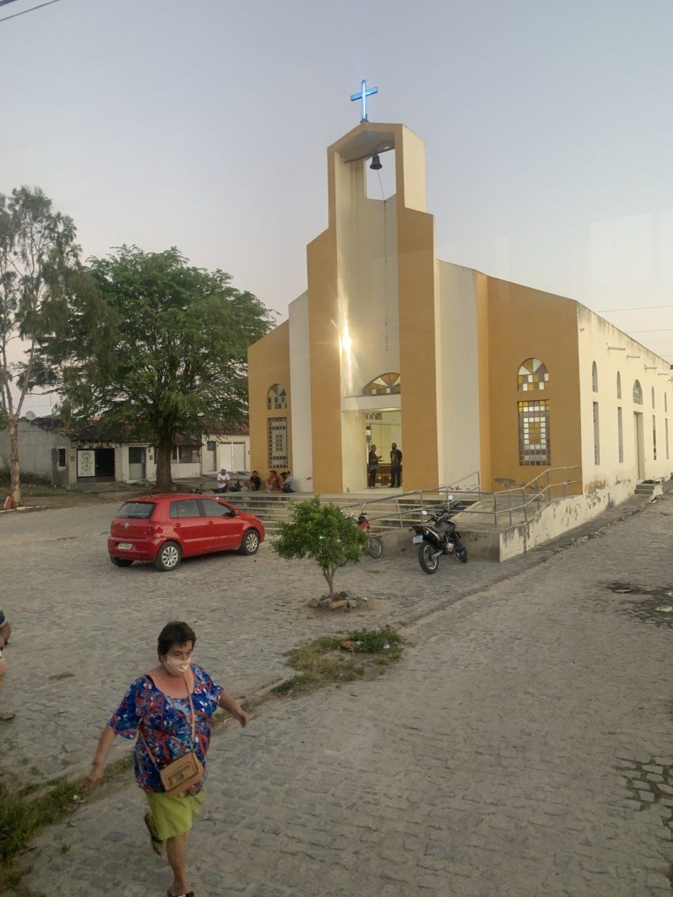 Mulher branca e de cabelo curto, vestindo blusa azul, bolsa de lado, máscara, em primeiro plano. Ao fundo, um carro vermelho, uma árvore grande e uma igreja. Sol fraco, provavelmente no entardecer.