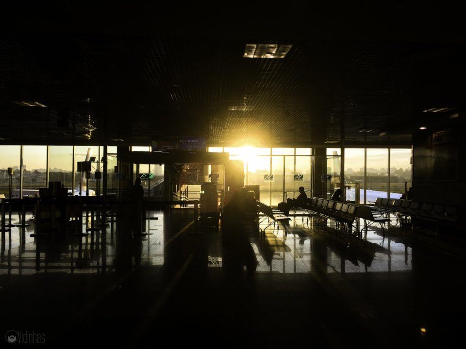 Foto de um terminal de aeroporto no entardecer, contra o Sol, revelando silhuetas de cadeiras e pessoas no saguão.