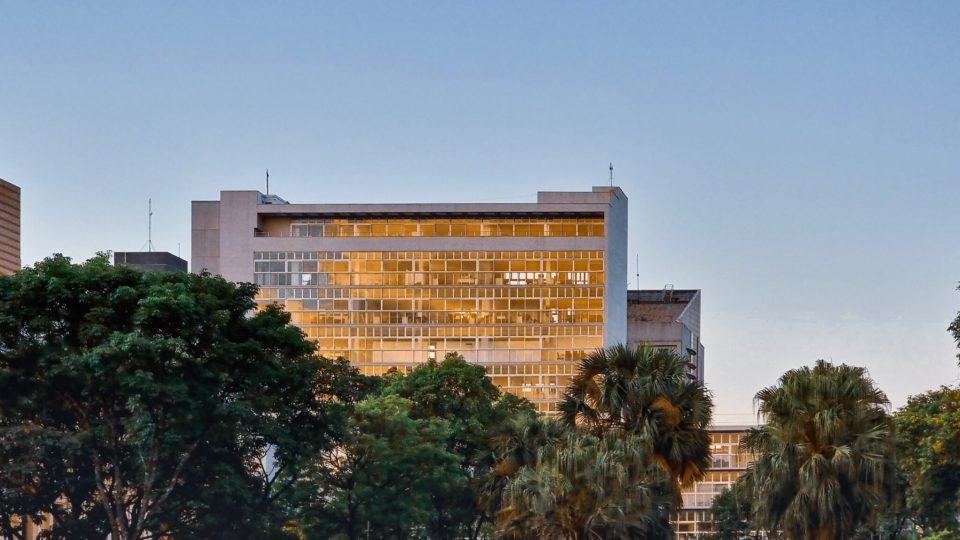 Prédio com muitas janelas, no entardecer, com as copas de várias árvores em primeiro plano.