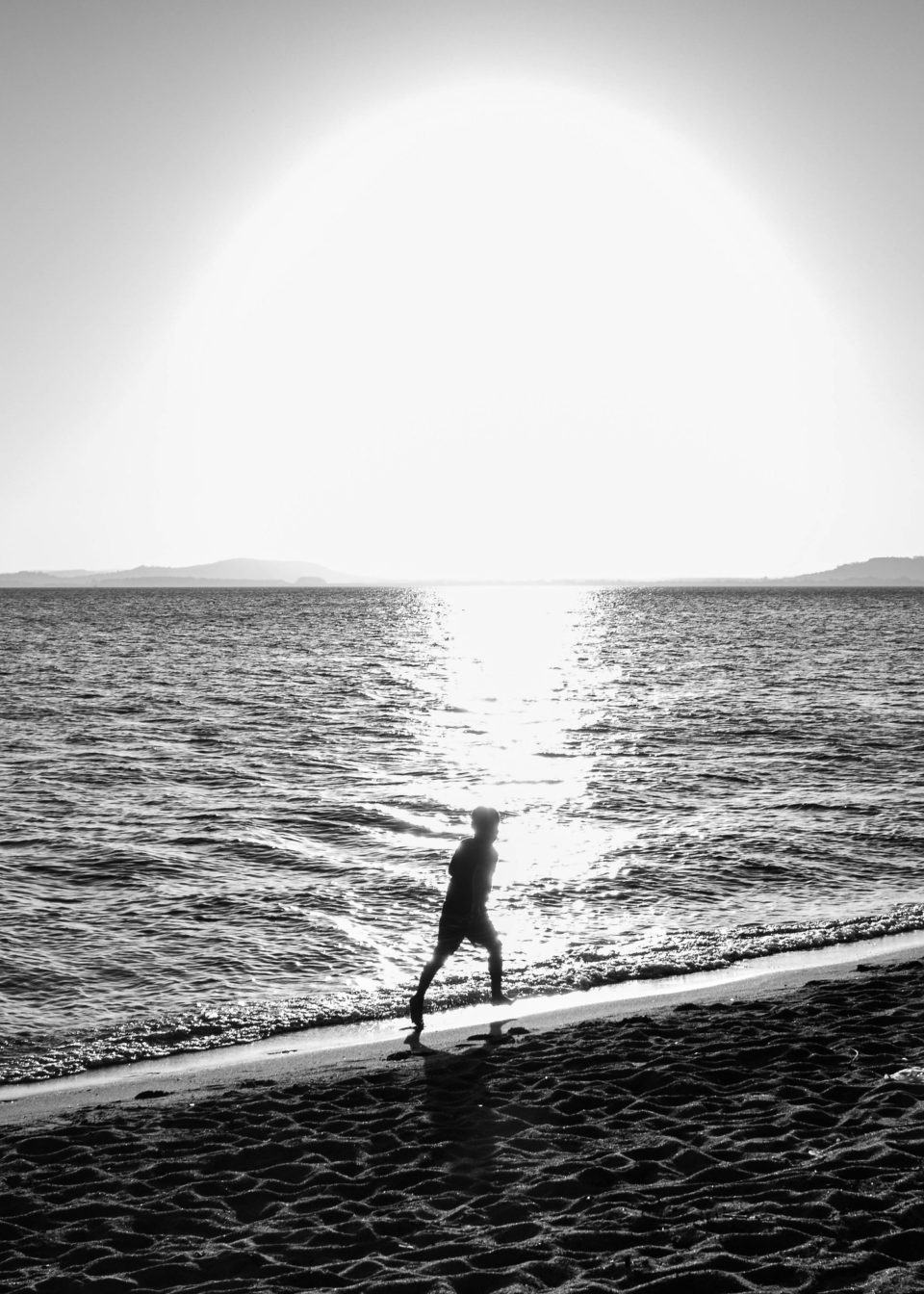 Foto de uma praia, com o mar e o Sol no horizonte, e a silhueta de uma pessoa caminhando à beira d'água.