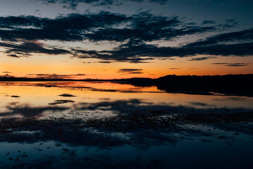 Entardecer, quase à noite. Foto de um lago/rio refletindo o céu com algumas nuvens escuras e uma parte, no horizonte, laranja/dourada.
