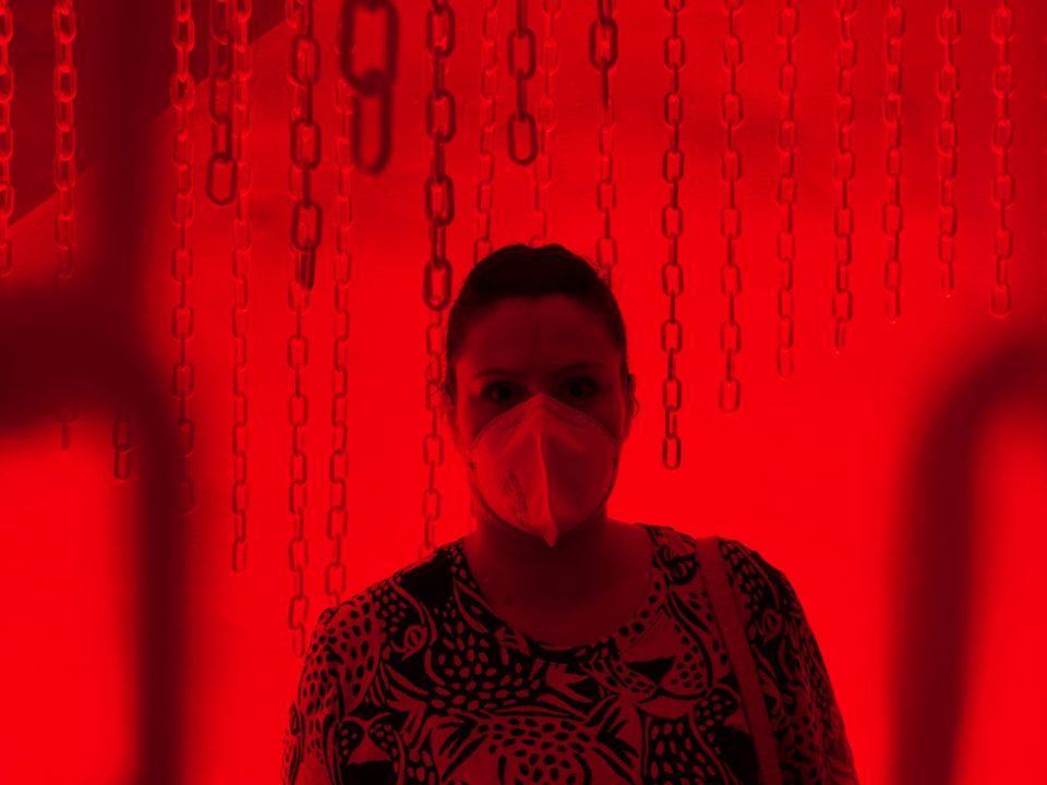 Mulher de máscara, em ambiente com luzes vermelhas bem fortes e correntes presas no teto.