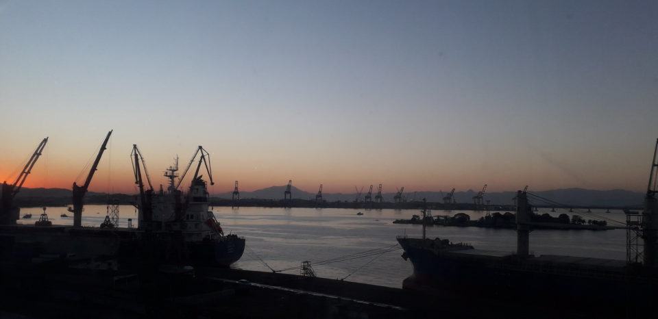 Foto do mar, no entardecer, com guindastes e embarcações quase indistinguíveis, parecendo somente suas silhuetas.