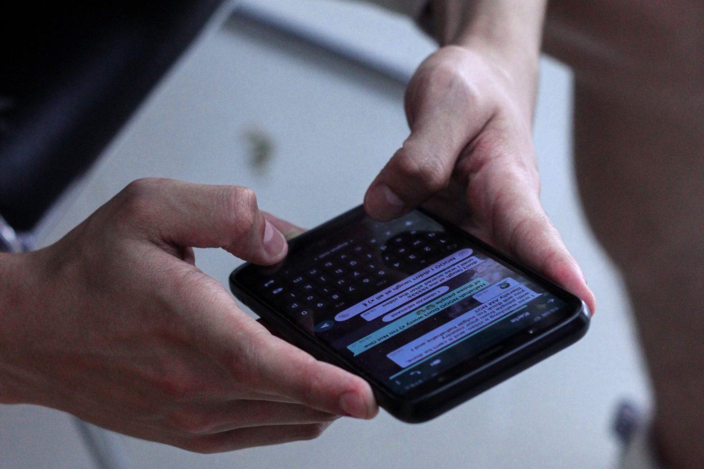 Visão de cima de duas mãos masculinas mexendo em um celular com uma conversa do WhatsApp (ilegível) aberta.