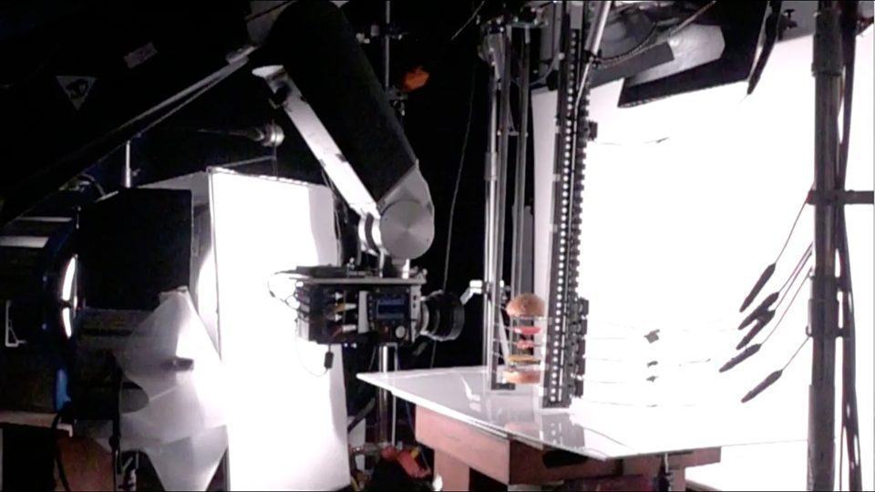 Foto de um braço robótico com uma câmera acoplada na ponta, apontando para uma estrutura com partes de um sanduíche sepradas, suspensas, contra um fundo branco.