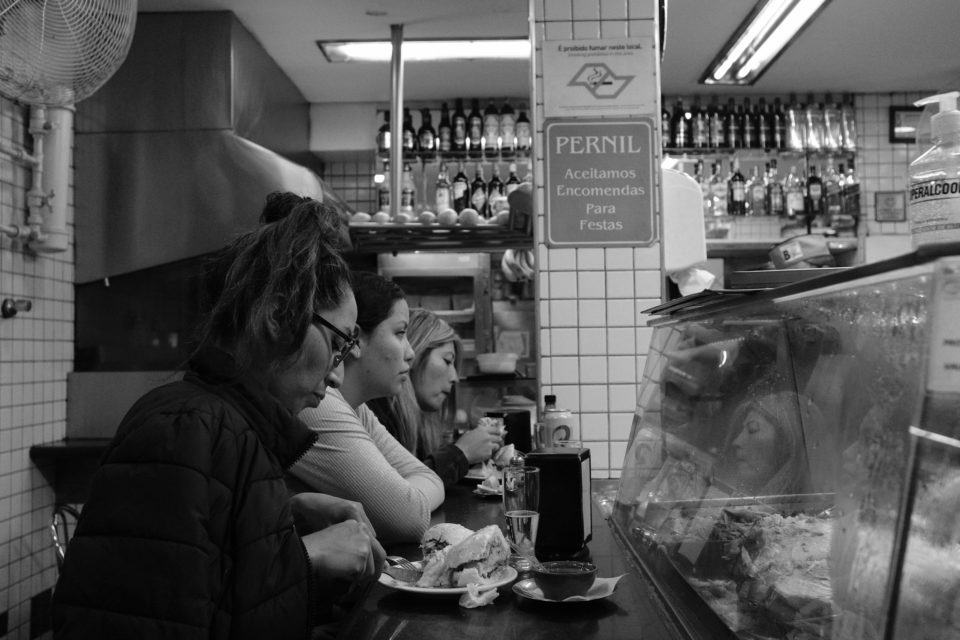 Foto em preto e branco de três mulheres encostadas em um balcão de padaria, de perfil, todas olhando para frente, as das pontas com pratos com comida. Ao fundo, uma pilastra com uma placa indicando a proibição de fumar em ambientes fechados e outra com os dizeres: “PERNIL. Aceitamos encomendas para festas”.