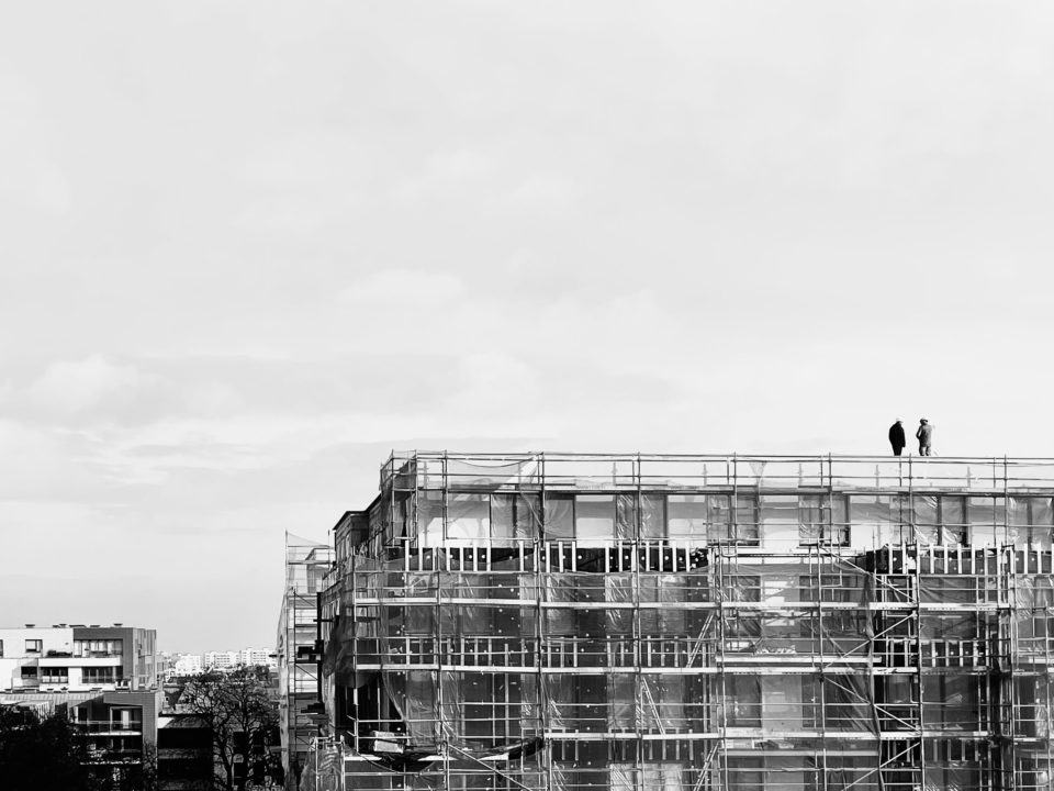 Foto em preto e branco de um prédio em construção, pegando bastante do céu. Em cima do prédio, duas pessoas.