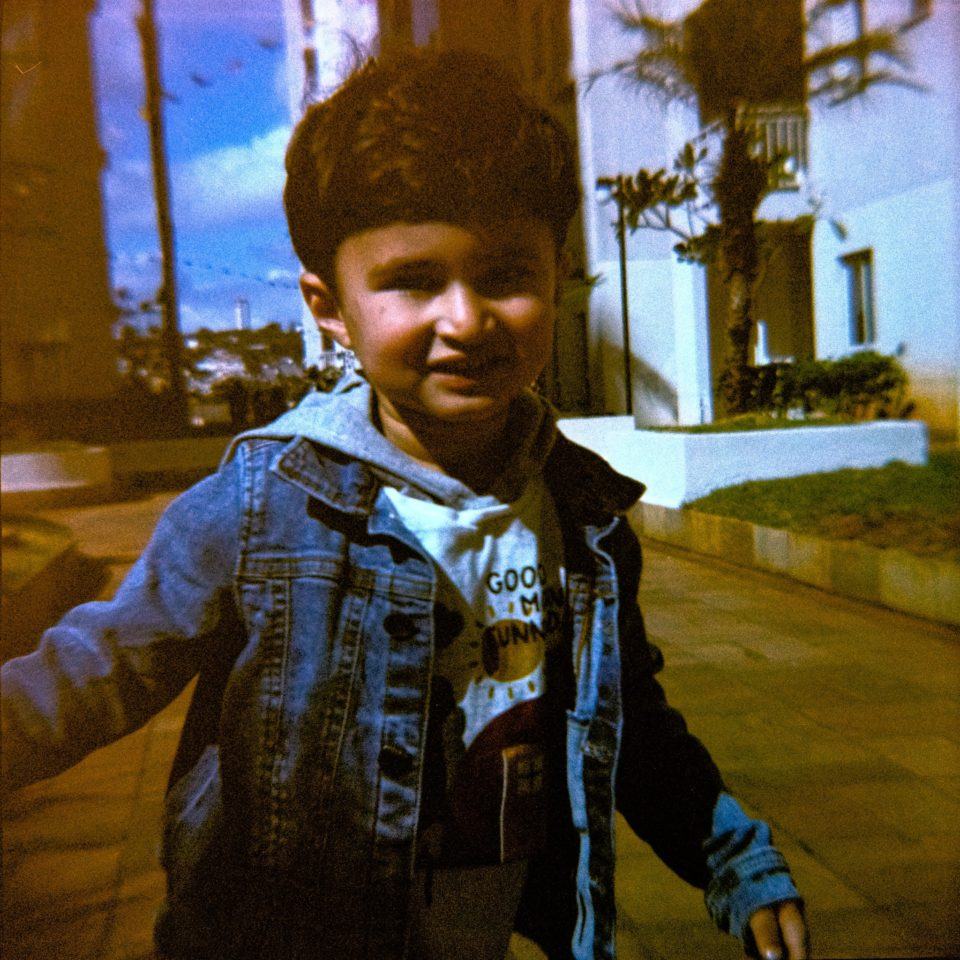 Menino sob o sol, vestindo jaqueta jeans, olhando para a câmera. Ao fundo, calçada e céu azul.