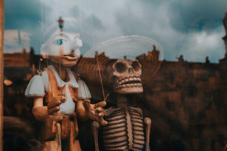 Dois bonecos, um pinóquio ao fundo e uma caveira com chapéu em primeiro plano, atrás de uma vitrine/vidro.