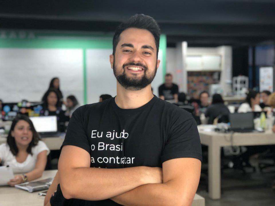 Homem branco, de barba e cabelo curto escuros, vestindo uma camiseta em que se lê “Eu ajudo o Brasil a contratar”, de braços cruzados, em um escritório.