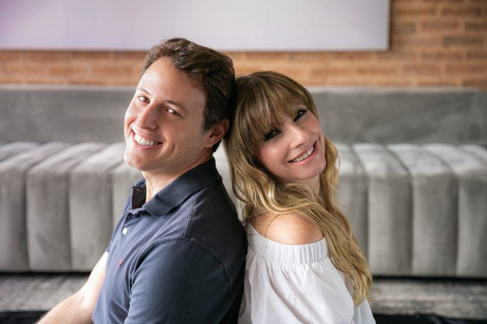 Homem e mulher, de perfil e de costas um para o outro, olhando para a câmera. Ambos brancos, ele de cabelo moreno, ela, loira. Ao fundo, um sofá.