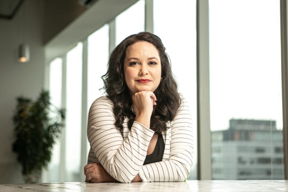 Mulher branca, de cabelos compridos e morenos, com os braços sobre uma bancada e a mão direita no queixo. Ao fundo, janelas grandes.