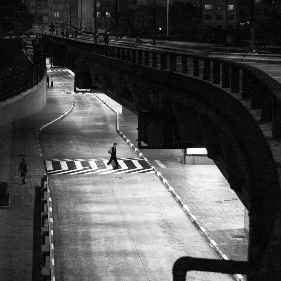 Foto em preto e branco de um homem cruzando uma faixa de pedestres à noite, bem iluminado, e o entorno escuro.