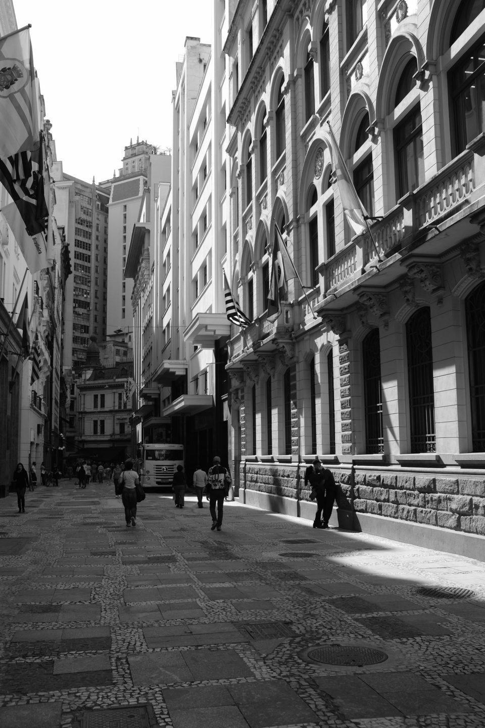 Foto em preto e branco de uma espécie de galeria a céu aberto, com o sol batendo de um lado e algumas pessoas transitando por ela.