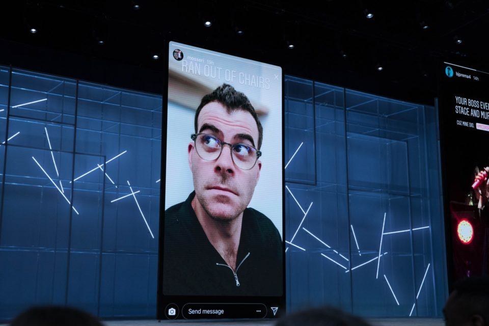 Homem branco, de cabelo escuro e óculos de grau, aparece no telão, enquadrado em uma tela de celular, olhando à sua direita. Ao fundo, palco azul escuro com iluminação baixa.