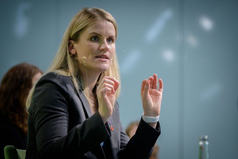 Mulher loira, de terninho cinza, com microfone no rosto e as duas mãos para cima, gesticulando.