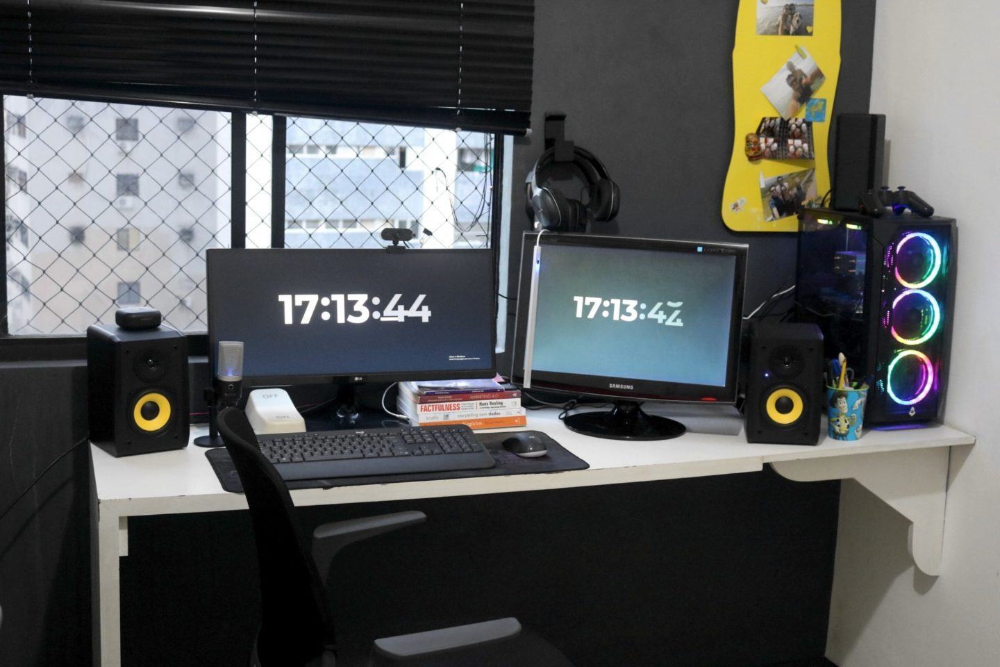 Mesa de trabalho branca, com dois monitores, acessórios e um desktop, todos pretos, e caixas de som pretas com detalhes amarelos. Ao fundo, parede e janelas pretas com detalhes amarelos.