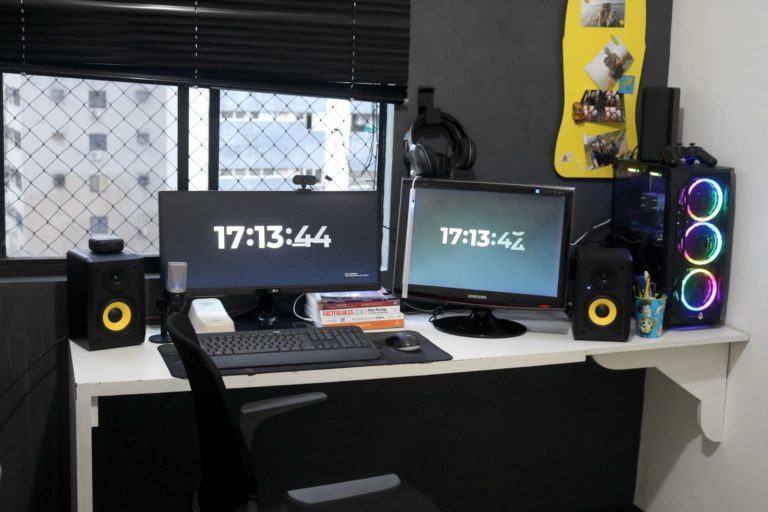 Mesa de trabalho branca, com dois monitores, acessórios e um desktop, todos pretos, e caixas de som pretas com detalhes amarelos. Ao fundo, parede e janelas pretas com detalhes amarelos.