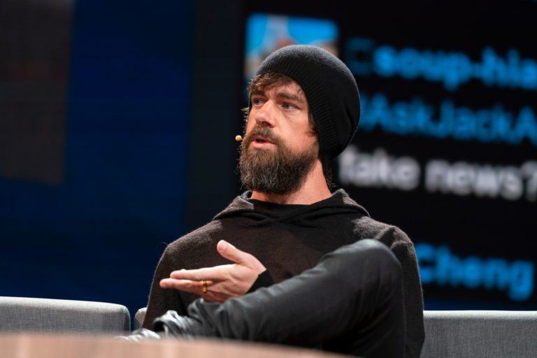 Foto levemente inclinada de homem branco, de barba escura comprida, roupa toda preta e gorro preto, gesticulando com a mão esquerda e falando a alguém à sua direita.